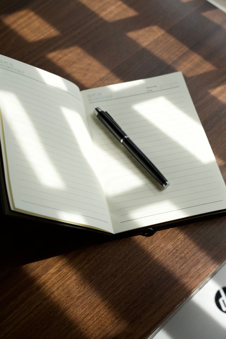 An open notebook and black pen on a wooden desk in sunlight.